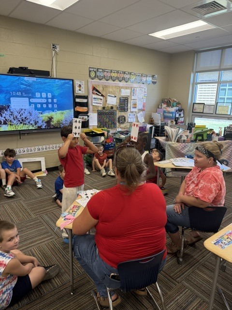 A classroom with a large screen on the wall. Several adults and children are seated. Some hold cards.
