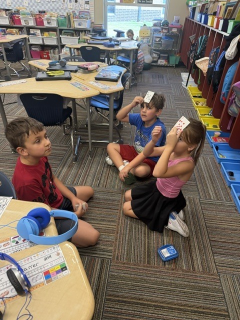 Three children seated on the floor in a classroom, wearing headphones. One holds a paper, and another looks at it.