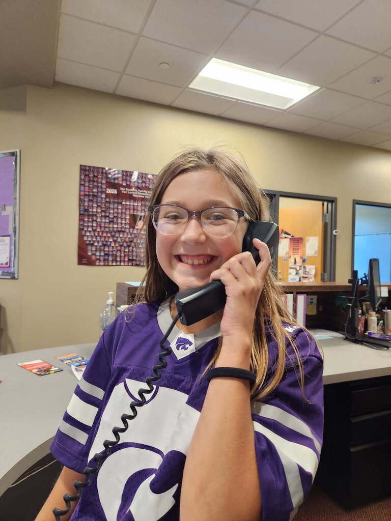 A girl in a purple shirt and glasses talks on a phone in an office with beige walls.