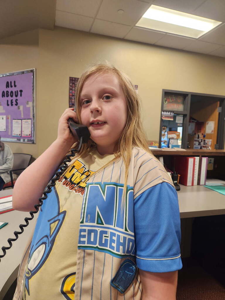 A child with blonde hair wearing a baseball jersey speaks into a phone. A bulletin board, books, and a desk are in the background.