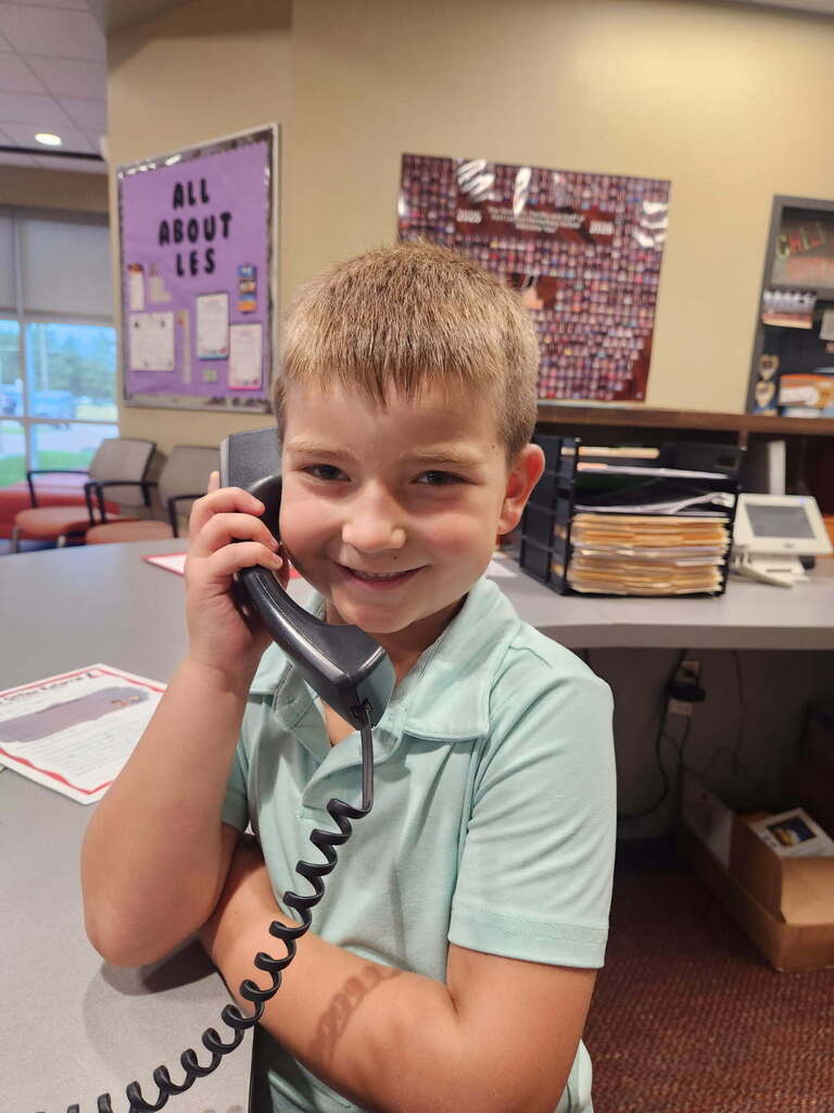 A young boy holds a phone to his ear, smiling. Behind him is a desk with various items.