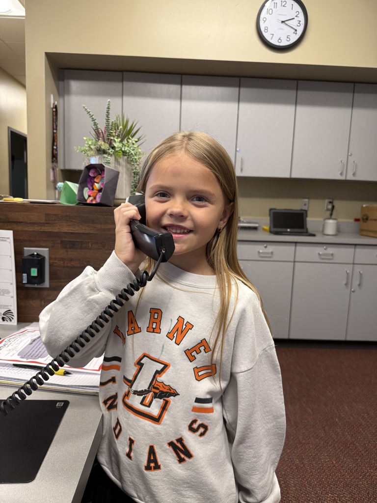 A young girl wearing a sweatshirt is talking on the phone in an office with white cabinets.