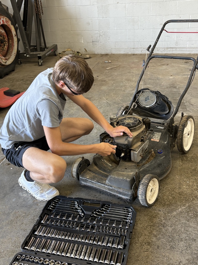 A young man kneels in a garage, working on a lawnmower. He wears a gray t-shirt, shorts, and glasses.