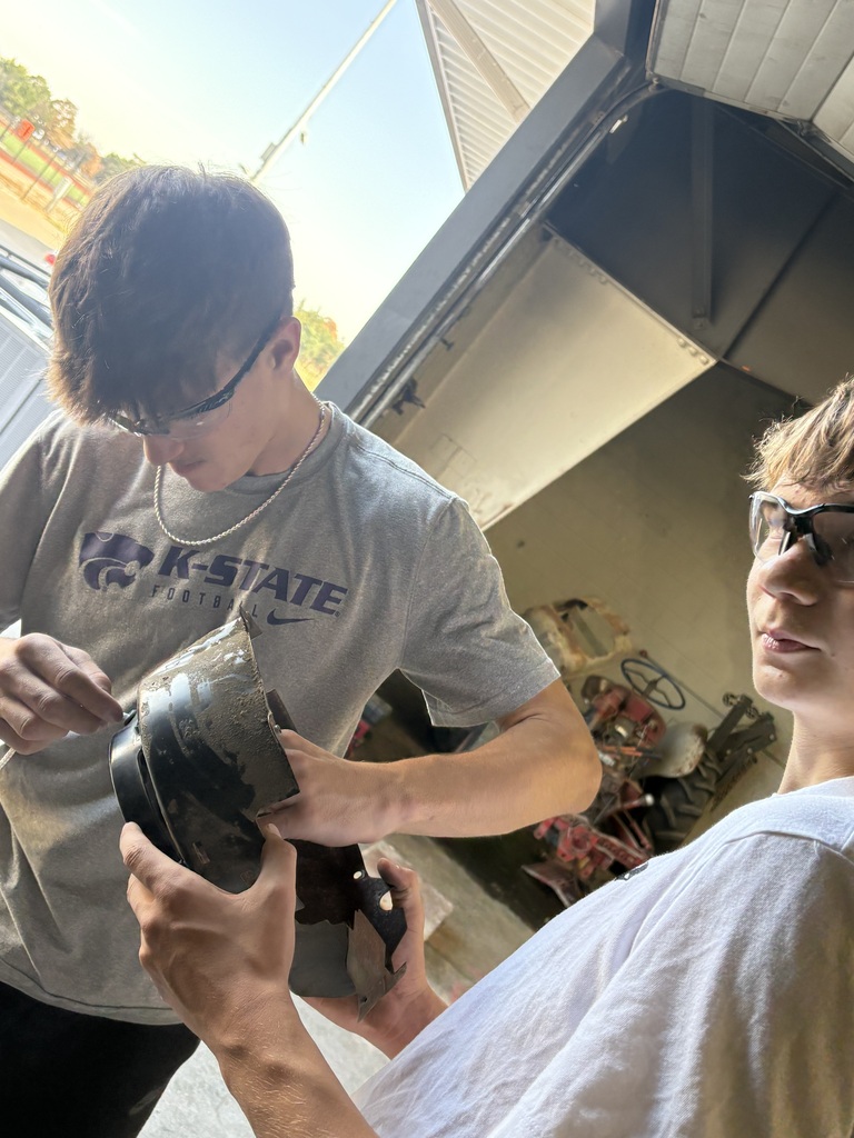 Two individuals wearing glasses are examining a piece of equipment. A garage with machinery in the background.
