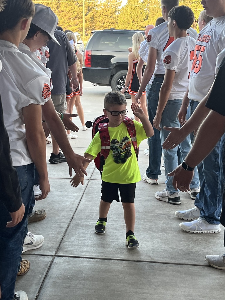 A group of individuals wearing school uniforms, some with backpacks, standing in a parking lot. A child in a green shirt walks through their hands.