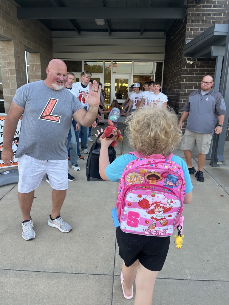 A man waves to a young girl in a pink backpack. They walk past a building with brick walls.