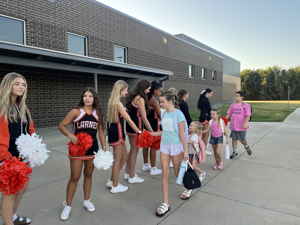 Cheerleaders in black, white, and red uniforms holding pom-poms stand by a building. Behind them, several children walk past.