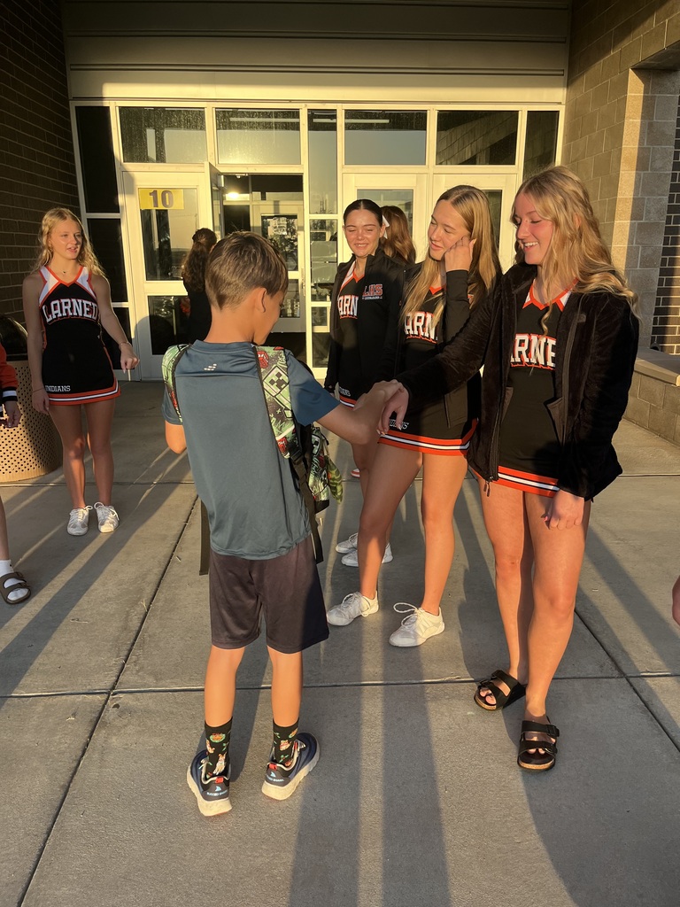 Four female students in sports uniforms greet a boy with backpacks. They stand on a sidewalk in front of a building.
