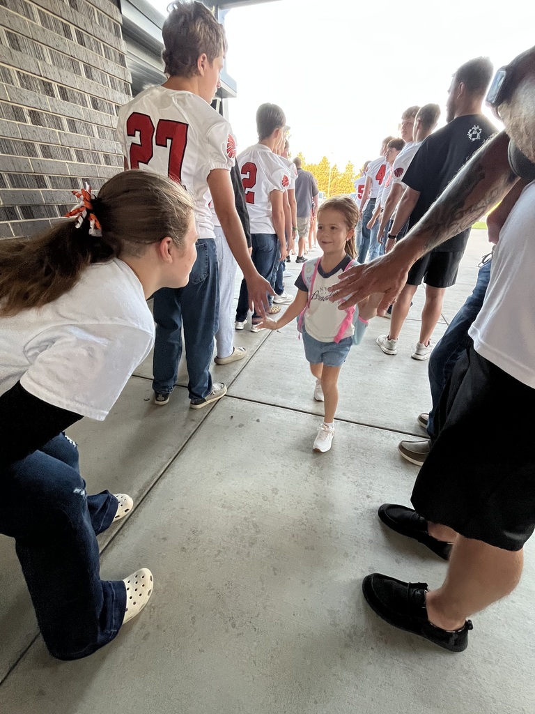 A group of adults and children in sports jerseys line up on a sidewalk with a brick wall in the background.