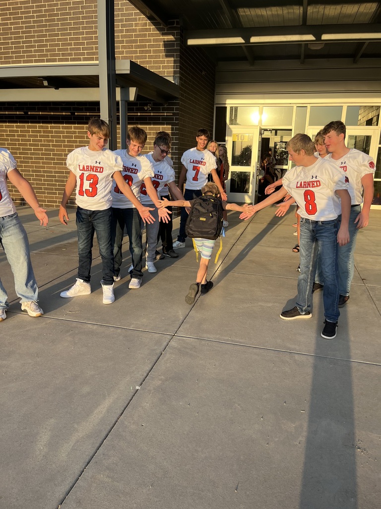 Nine individuals, including a young child, wear football jerseys with numbers and letters. They are walking in front of a building.