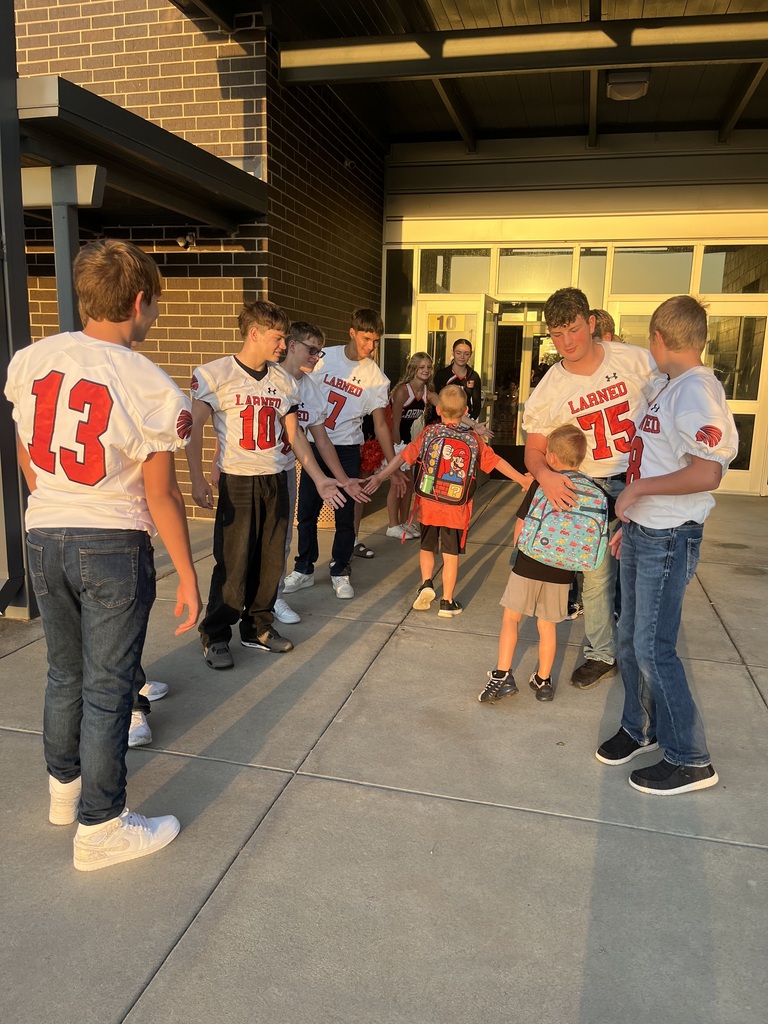 Several adults and children, some in football jerseys, stand on a concrete sidewalk in front of a building.