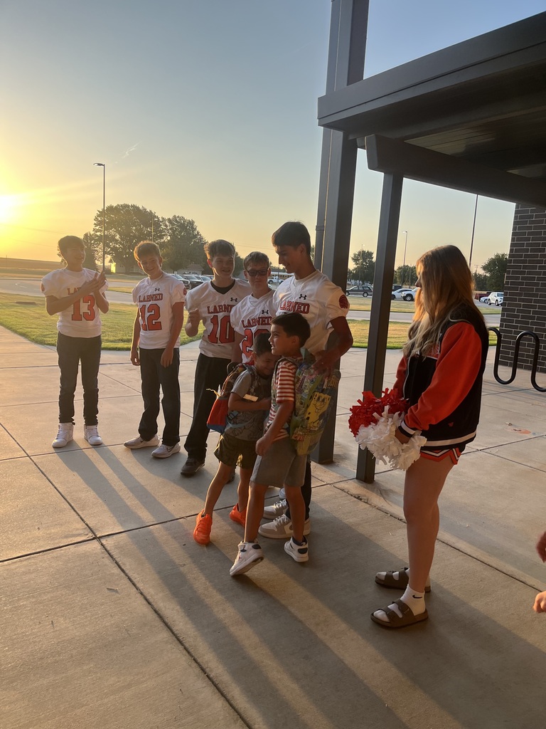 A group of people stands outside a building at sunset. Some wear jerseys, and one person holds flowers.