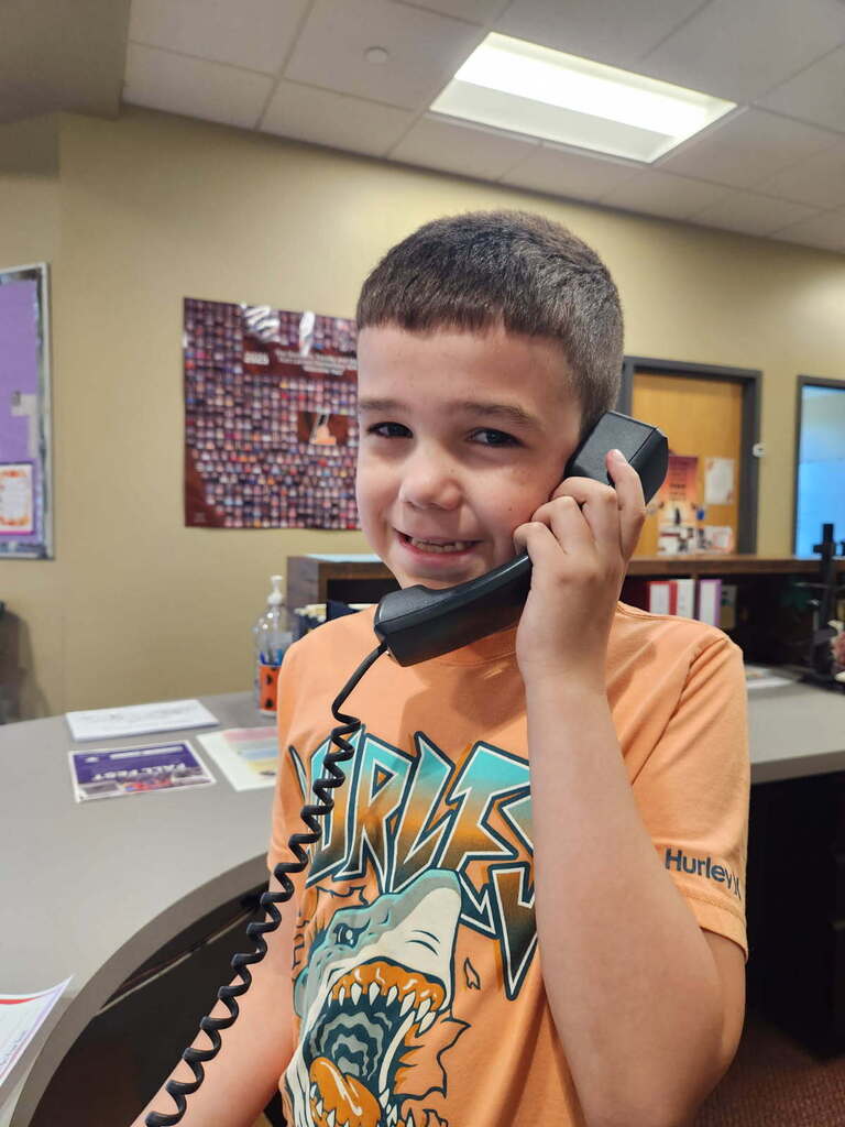 A boy wearing an orange shirt with a shark design smiles and talks on a phone. Behind him is a beige wall with a large picture.