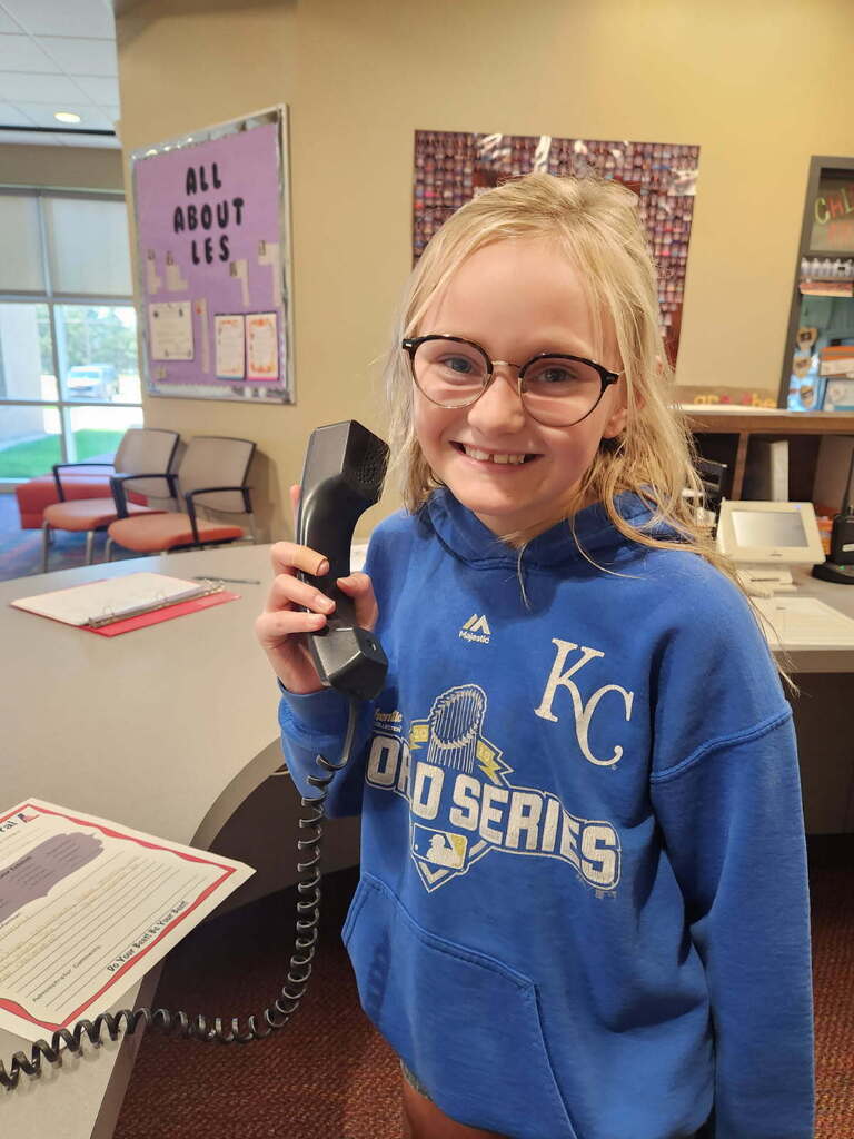 Blonde girl with glasses holds a telephone, smiling, in a room with a desk, chairs, and a bulletin board.