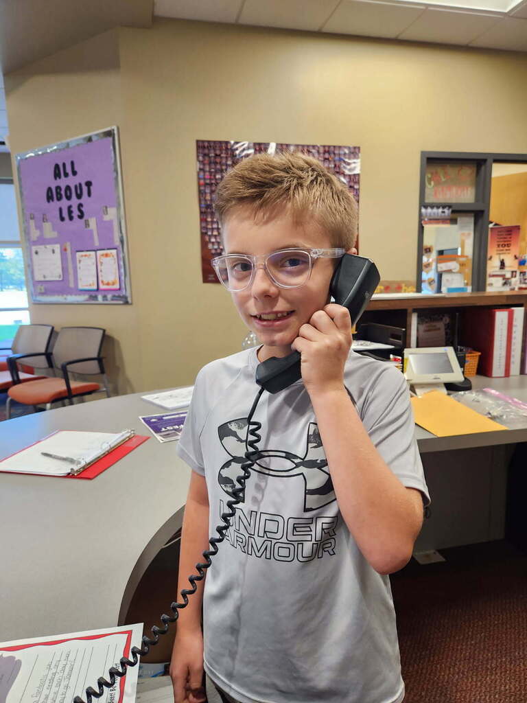 A young boy wearing glasses stands in an office with a desk and chairs in the background.