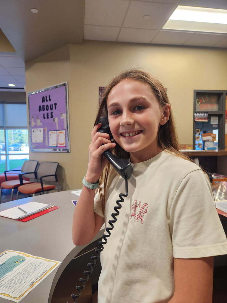 A young girl stands at a desk in a classroom, holding a phone to her ear and smiling.