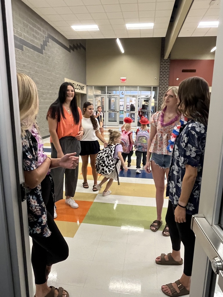 Five adults and a child stand in a hallway with multicolored flooring. People in the background wear hats and carry backpacks.