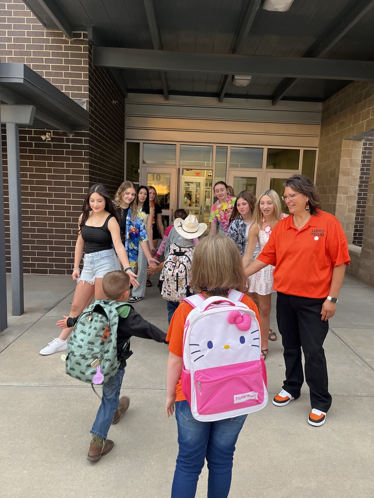 A group of children and adults are outdoors in front of a building. Some are carrying backpacks.