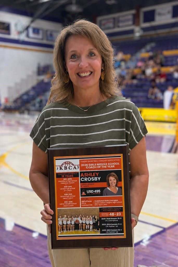 A woman holds a plaque with a team photo, standing in a sports arena. The plaque reads "Ashley Crosby".