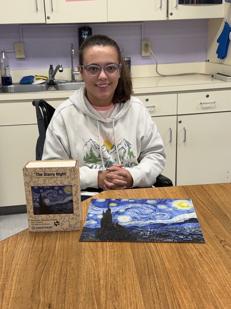 A person sits at a table with a puzzle and book, wearing glasses, in a kitchen.