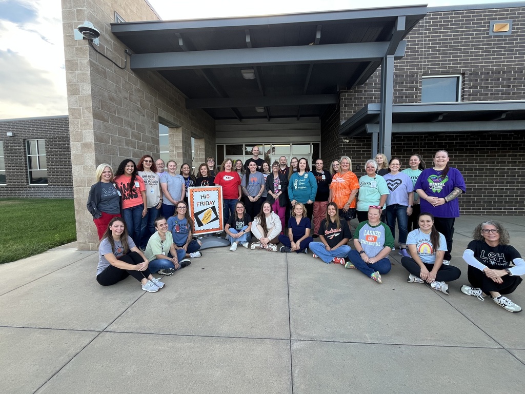 A group of people are gathered outside a brick building, posing for a photo on a sidewalk.