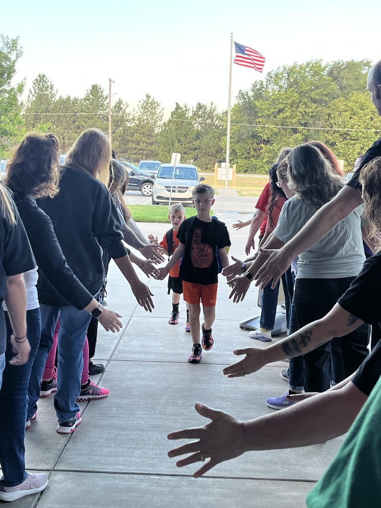 A group of adults and children stand in a circle, hands linked, on a sidewalk. An American flag is visible in the background.