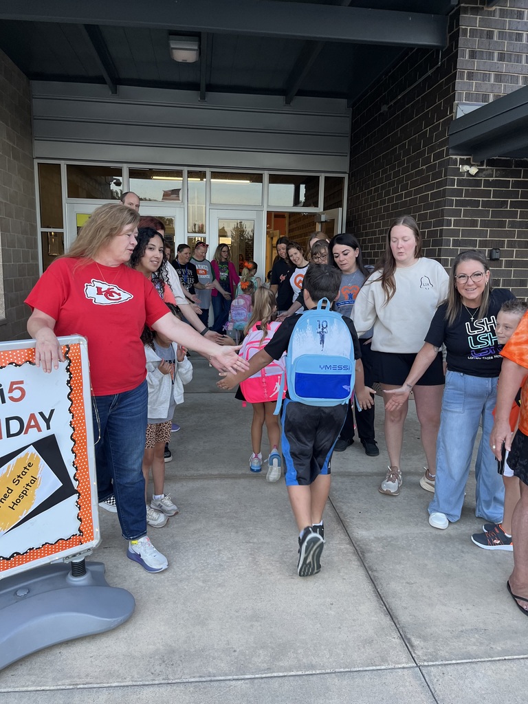 People stand in line outside a building with glass doors. A woman in red holds a sign, a child in a backpack walks ahead.
