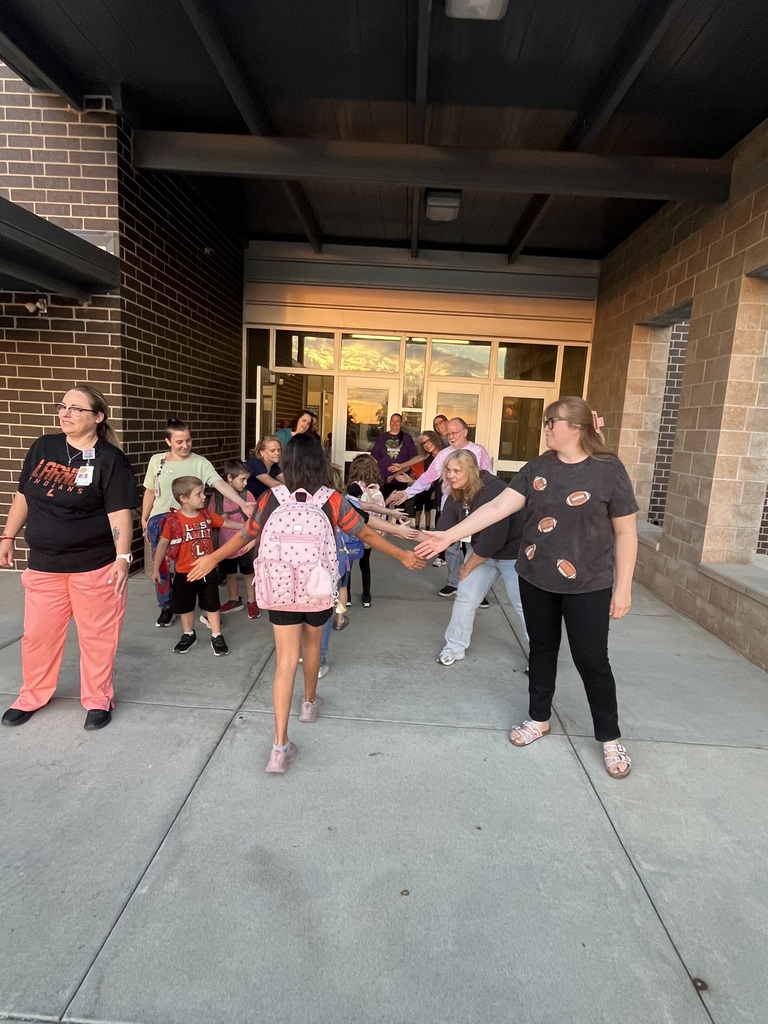 Adults and children hold hands in a circle. A brick building with glass doors and windows is in the background.