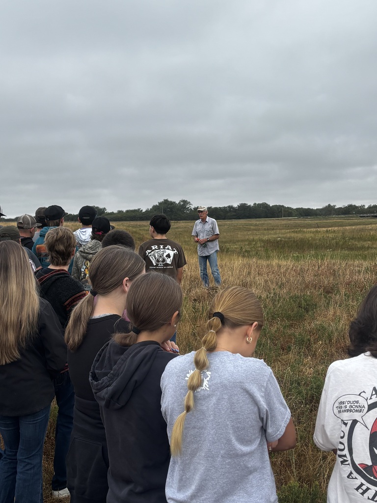 A group of students outdoors in a grassy field, listening to a person standing in front of them.