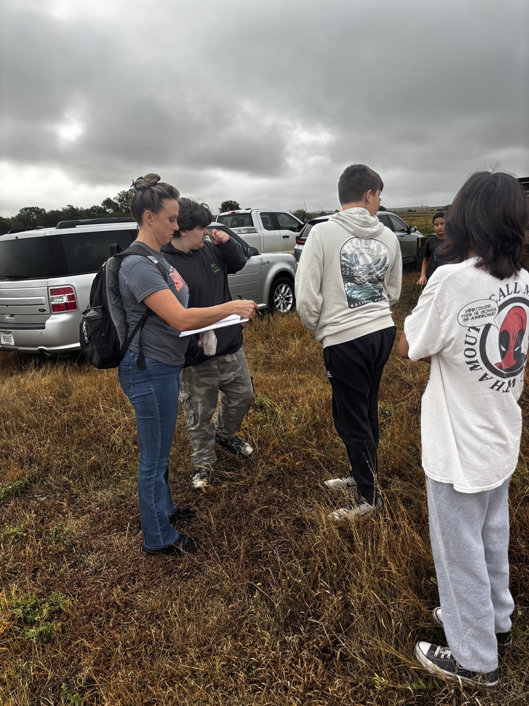 A group of people standing in a field. One holds a paper. Vehicles are parked in the background.