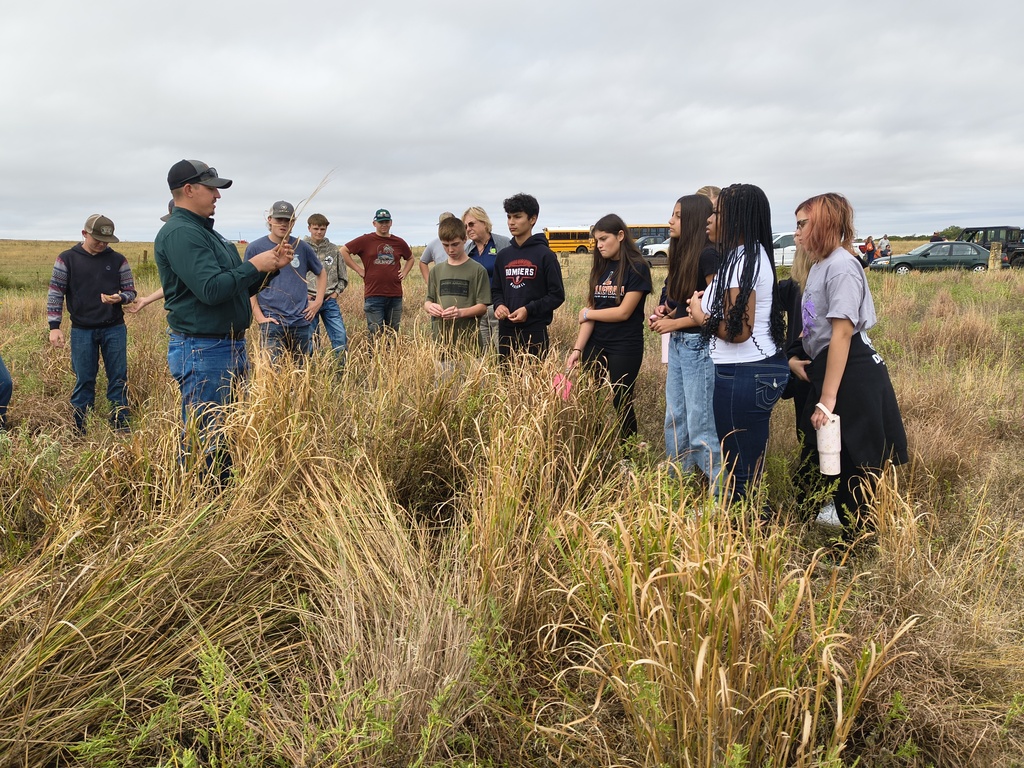 A group of people stands in a field with tall grass. One person, wearing a hat and green jacket, leads the group.