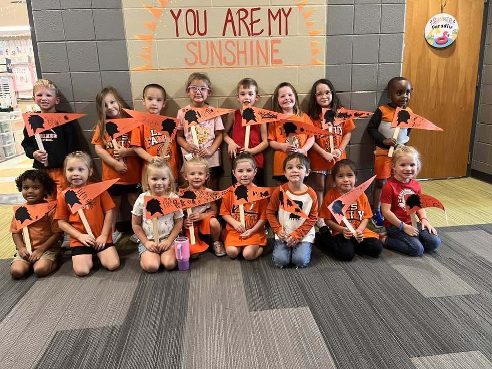 Group of children in orange shirts holding orange flags, standing in a line in a room.