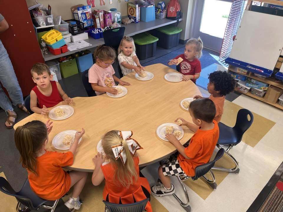Preschool children seated around a table, each holding a plate, in a classroom with storage boxes.