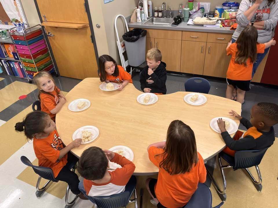 Children in orange shirts sit around a table eating. A woman stands behind the table in a kitchen.