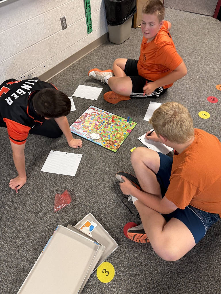 Three boys are playing a board game on the floor. They sit on the floor and have papers and game pieces around them.