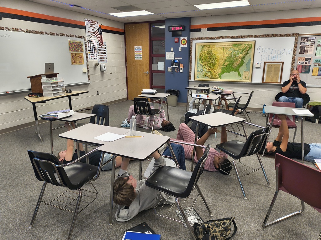 A classroom with desks arranged in rows, students lying on the floor, a teacher at a desk, and a map on the wall.
