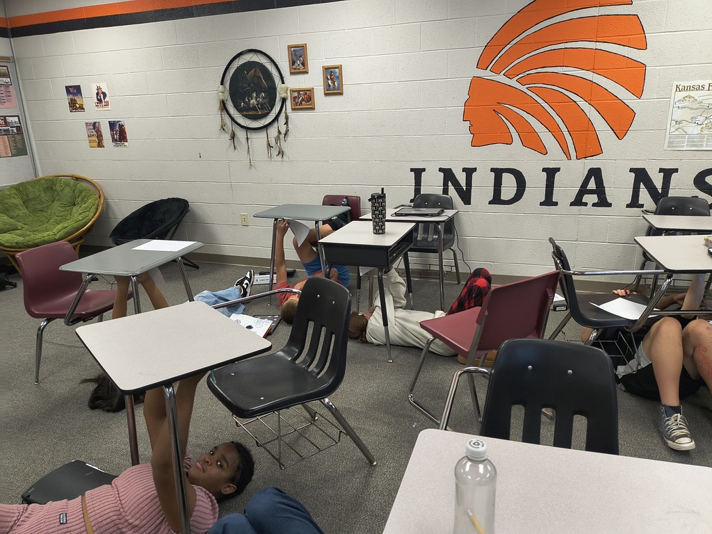 Classroom with desks and chairs, walls with posters and a large orange logo reading "INDIANS". Students are seated on the floor.