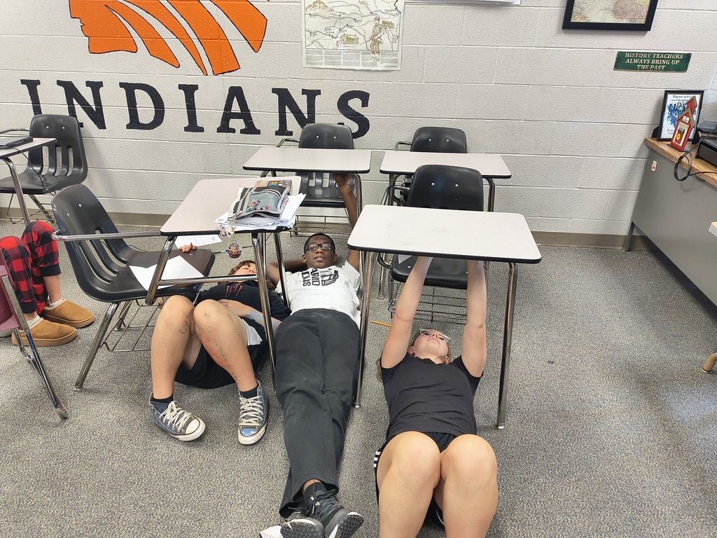 A classroom with desks, chairs, and three students lying on the floor. The wall reads "INDIANS".