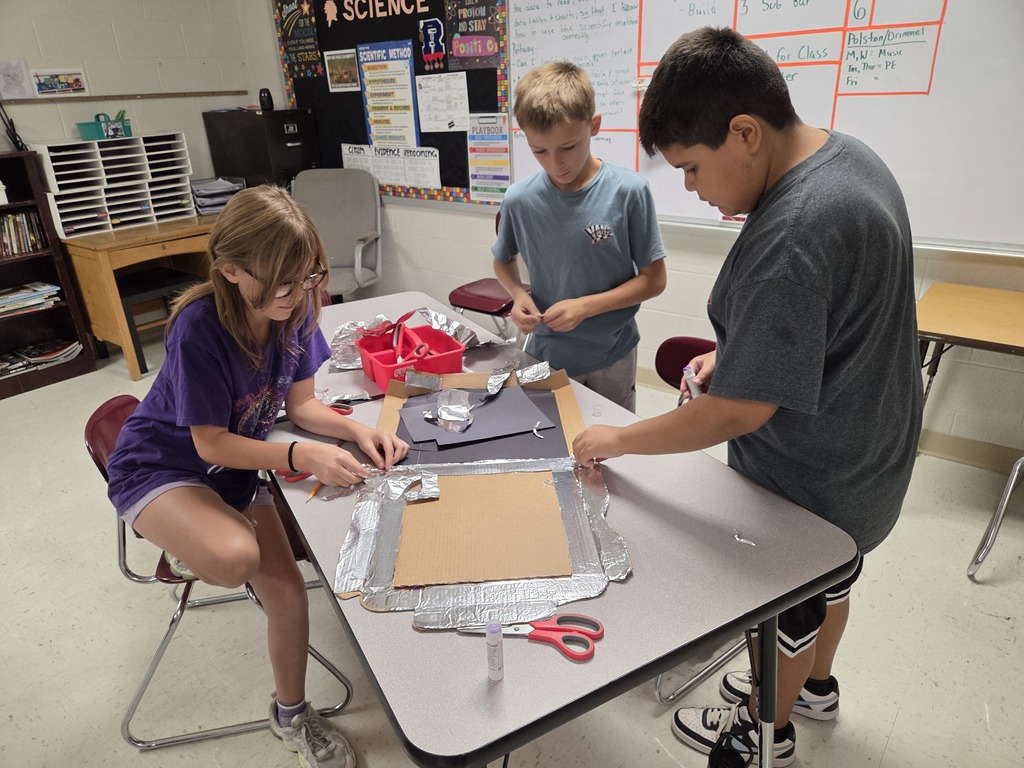 Three students collaborate on a science project at a table, with materials like paper and scissors.