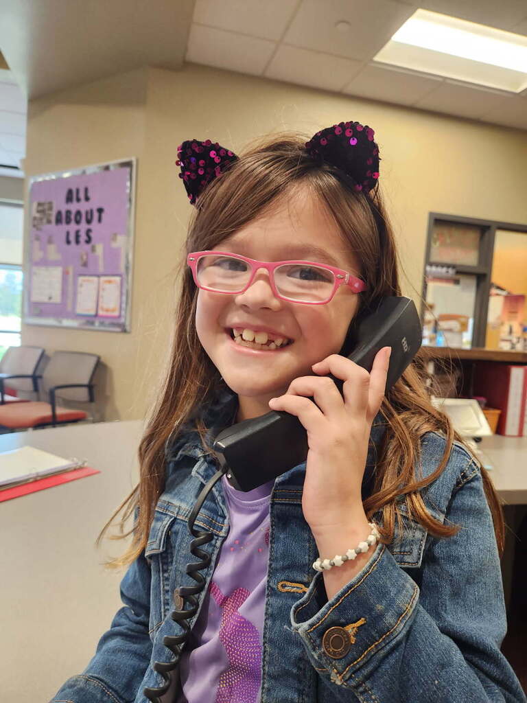 A young girl with pink glasses and cat ears sits in a classroom. She holds a telephone to her ear and smiles.