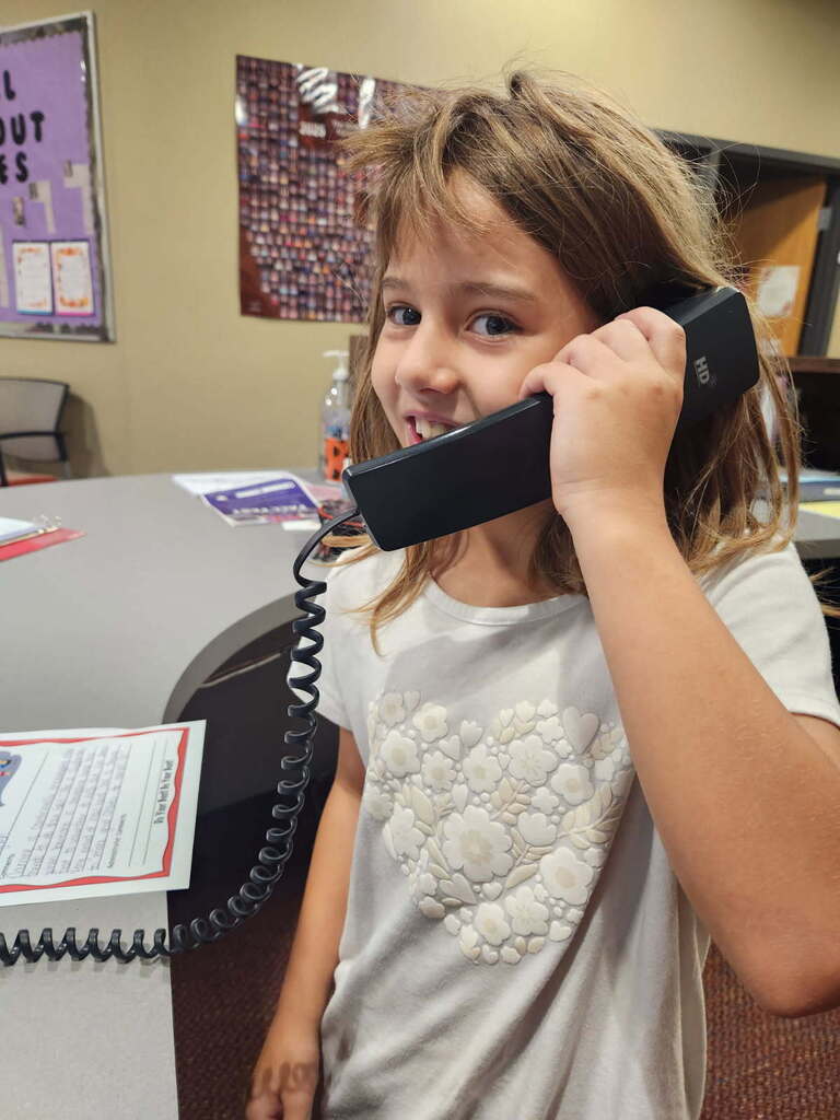 A young girl holds a phone to her ear, smiling while standing in a room. Behind her, there is a desk with papers and a bulletin board.