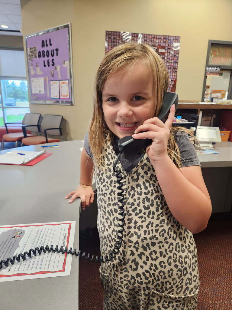 A young girl in a leopard-print outfit stands by a desk, holding a phone to her ear and smiling.
