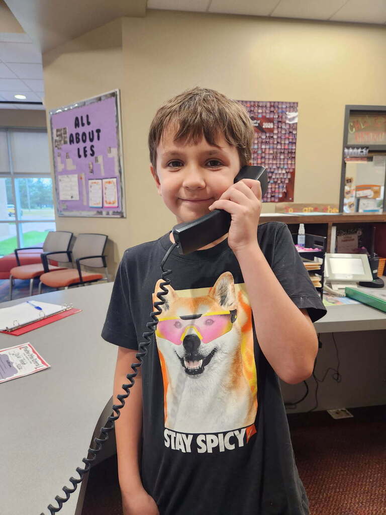 A boy in a black shirt stands in an office, holding a black corded phone to his ear.