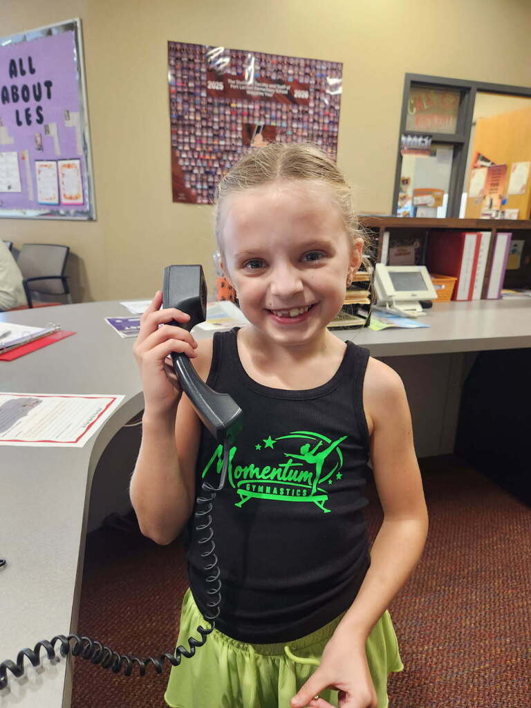 A young girl with blonde hair holds a telephone to her ear. She wears a black tank top and green skirt. Behind her is a desk with papers and a bulletin board.
