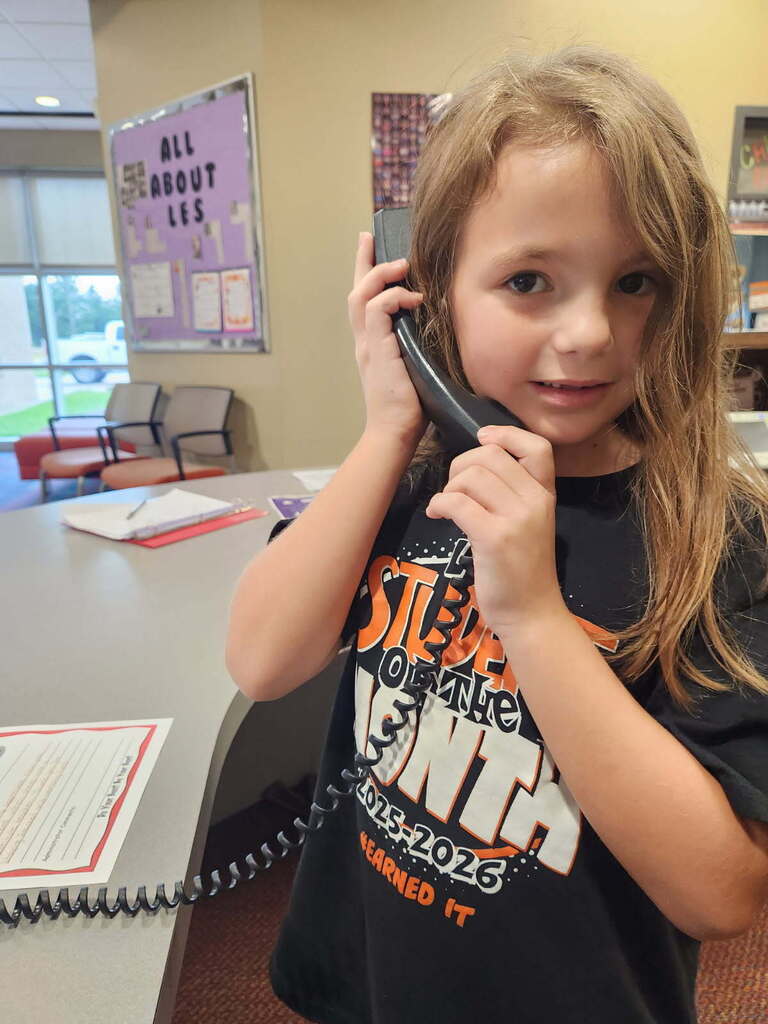 A young girl stands in an office, holding a phone to her ear. She wears a T-shirt with a sports team logo.