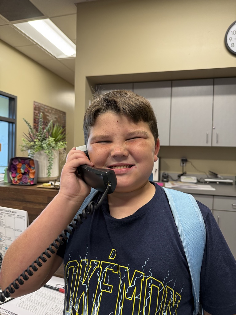 A young boy is smiling and holding a telephone to his ear. He is wearing a blue t-shirt with printed text and has a backpack on his back.