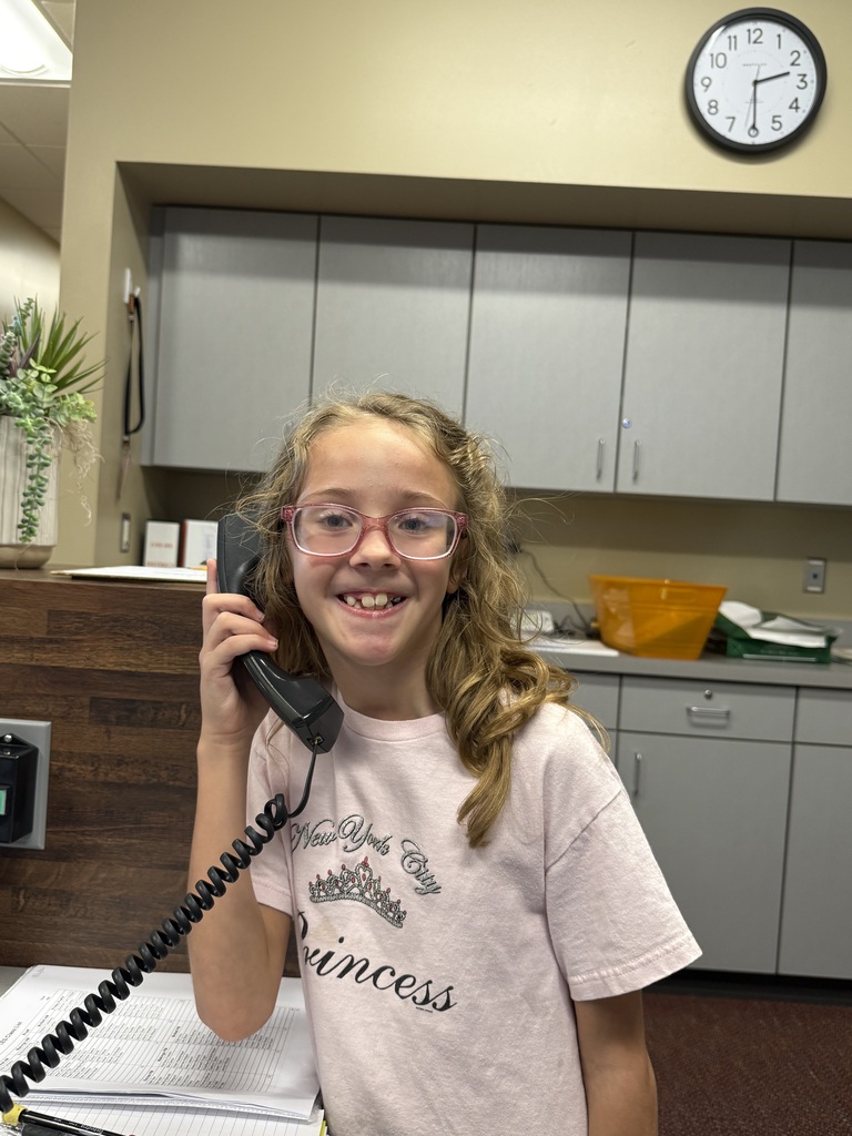 A young girl in glasses stands in an office, holding a phone, smiling, with cabinets and a plant behind her.