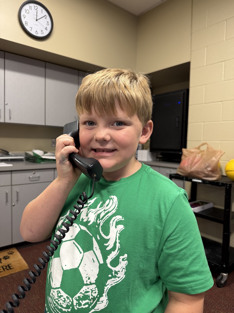 A boy in a green t-shirt smiles while talking on a phone in a room with cabinets and a clock.