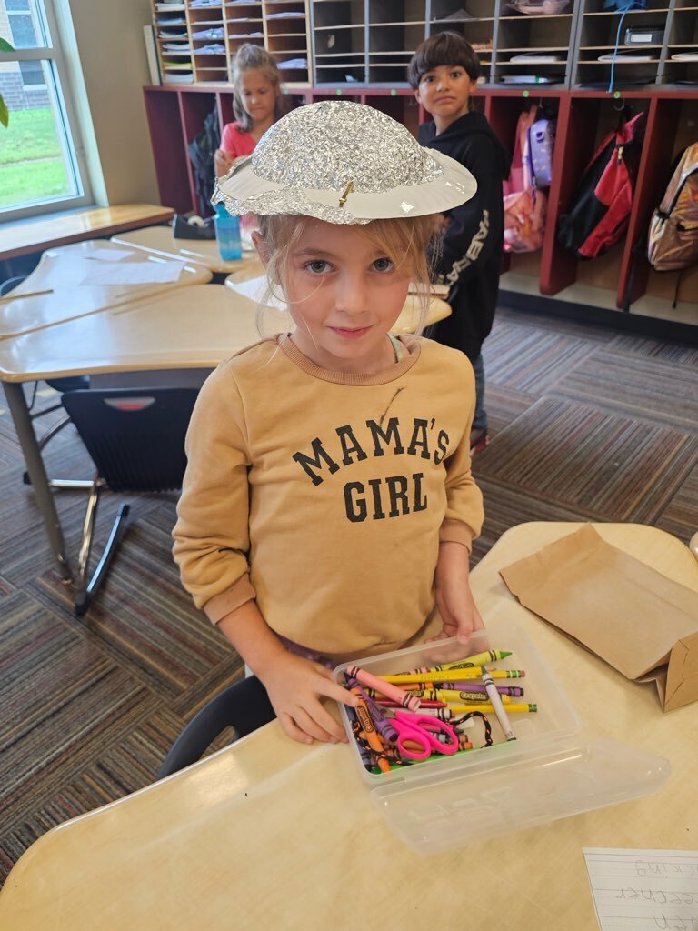 Child wearing a hat and holding a plastic container of crayons, sitting at a desk.
