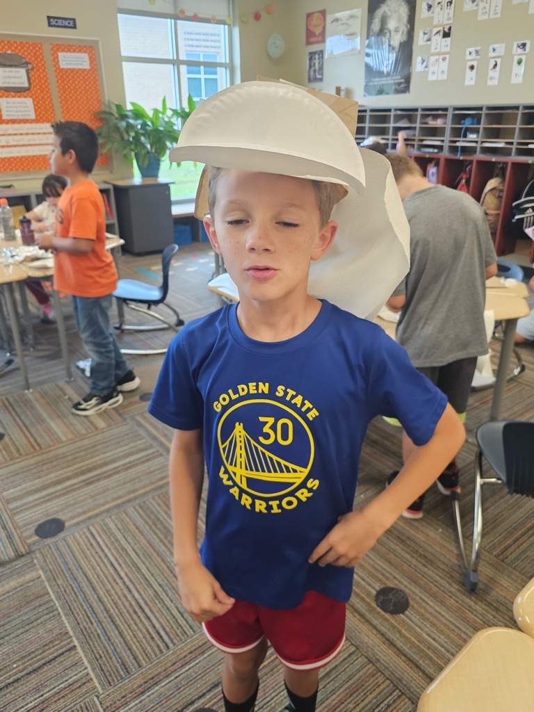 Boy wearing a blue Golden State Warriors shirt, red shorts, and a paper plate hat stands in a classroom.
